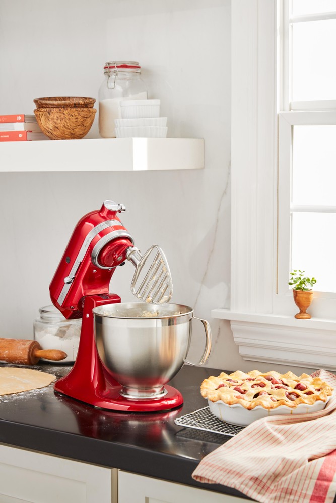 Red KitchenAid stand mixer with a stainless steel bowl and pastry beater attachment, positioned on a black countertop beside a pie and baking tools.