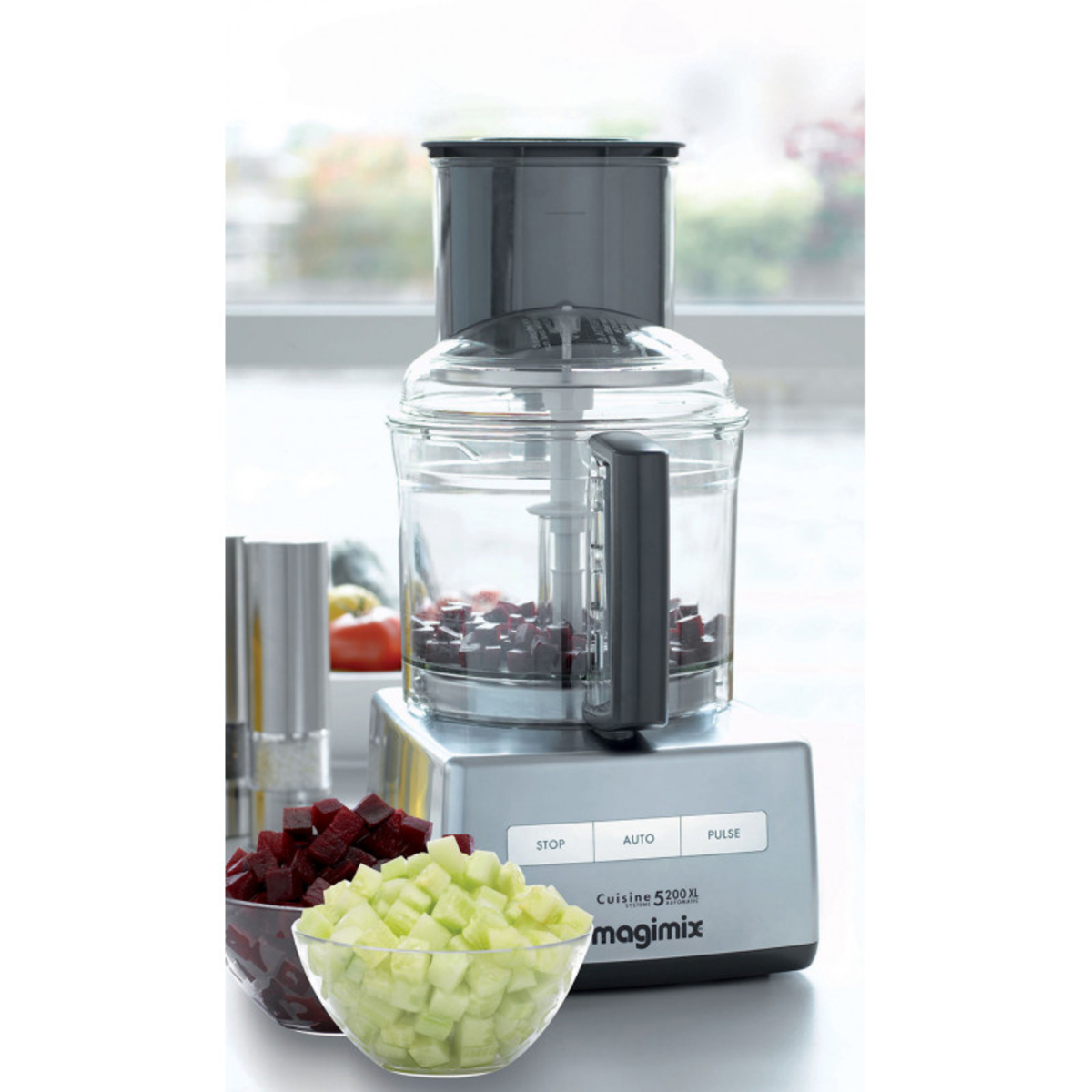 Satin silver Magimix 5200XL food processor with a transparent bowl, black lid, and control panel, accompanied by chopped vegetables in bowls.