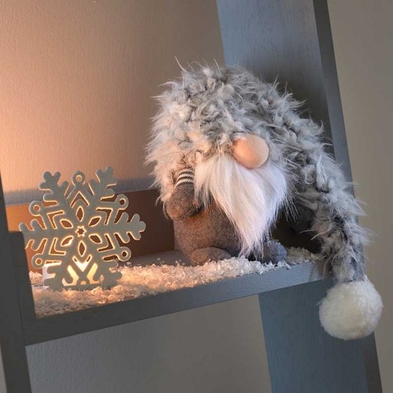 Grey plush winter gnome with a fluffy hat and white beard, sitting beside a decorative snowflake light on a shelf.