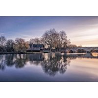 Countryside hotel by a river with arched stone bridge, surrounded by trees under a pastel sky.