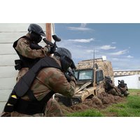 Paintball players in camouflage gear and helmets aiming markers near a camouflaged vehicle on grassy field.