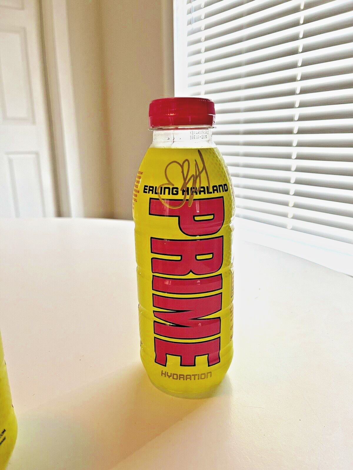 Bright yellow plastic bottle of Prime Hydration drink featuring Erling Haaland's name, a red cap, and bold pink lettering, part of a multipack of 12 x 500 ml bottles.