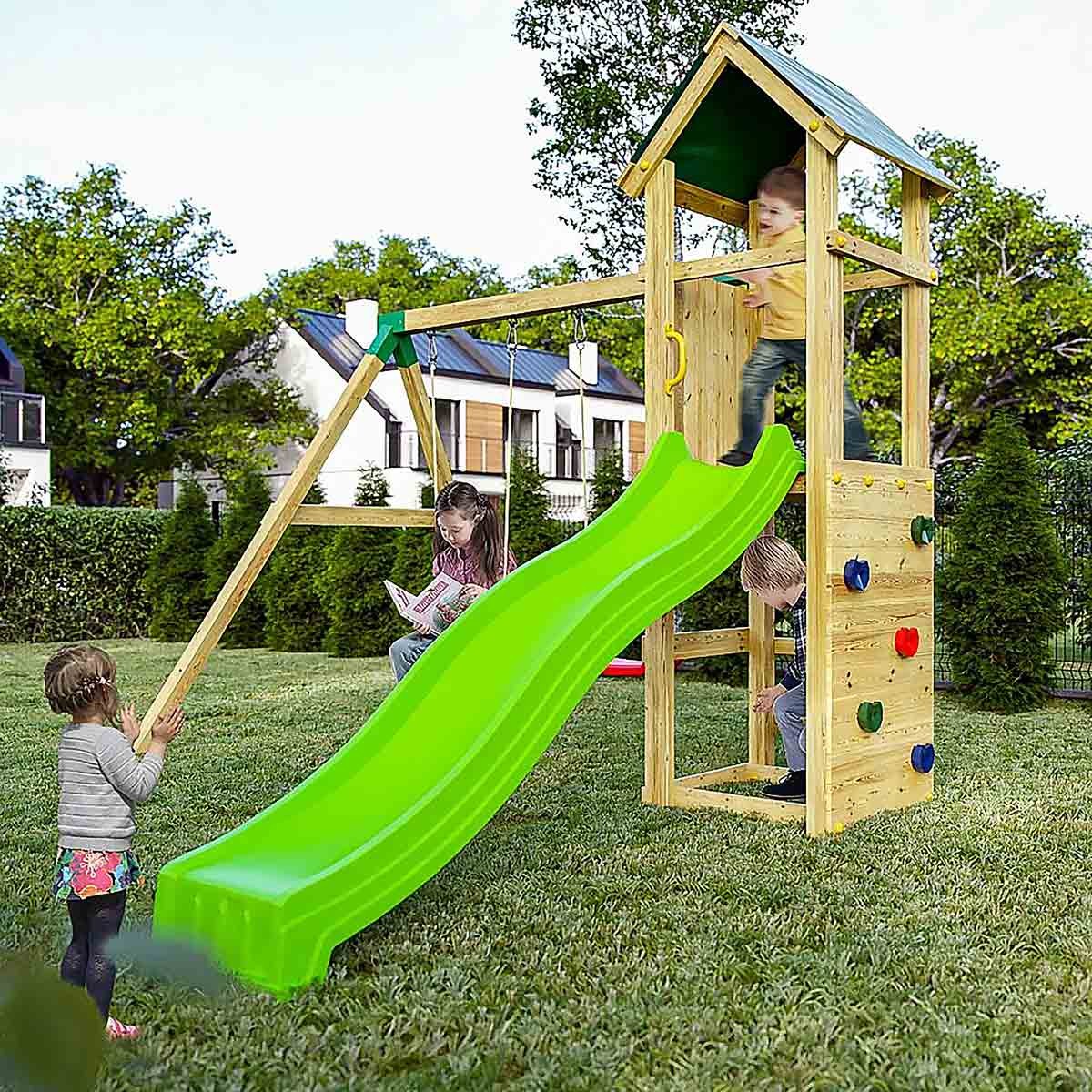 Brown wooden play fort with a green slide, climbing wall, swings, and a small roof, set in a grassy area with children playing around it.