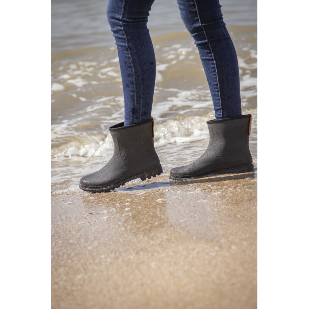 Black waterproof women's riding boots with a textured surface and a low ankle design, paired with blue jeans, standing on a sandy beach near the water.