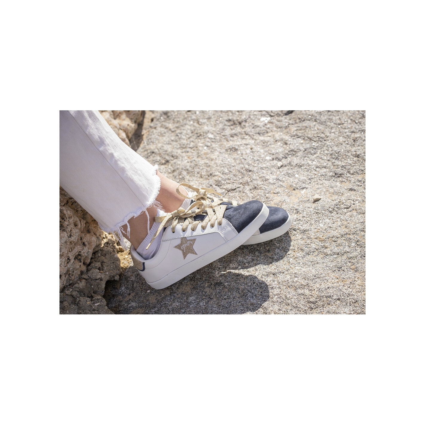 White and navy suede women's trainers by Pénélope, featuring a gold star detail and beige laces, styled with light-colored pants on a rocky surface.