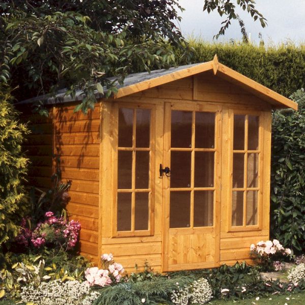 Traditional wooden summer house in natural wood finish, featuring double glass doors, large front windows, and a sloped roof, surrounded by colorful flowers and greenery.
