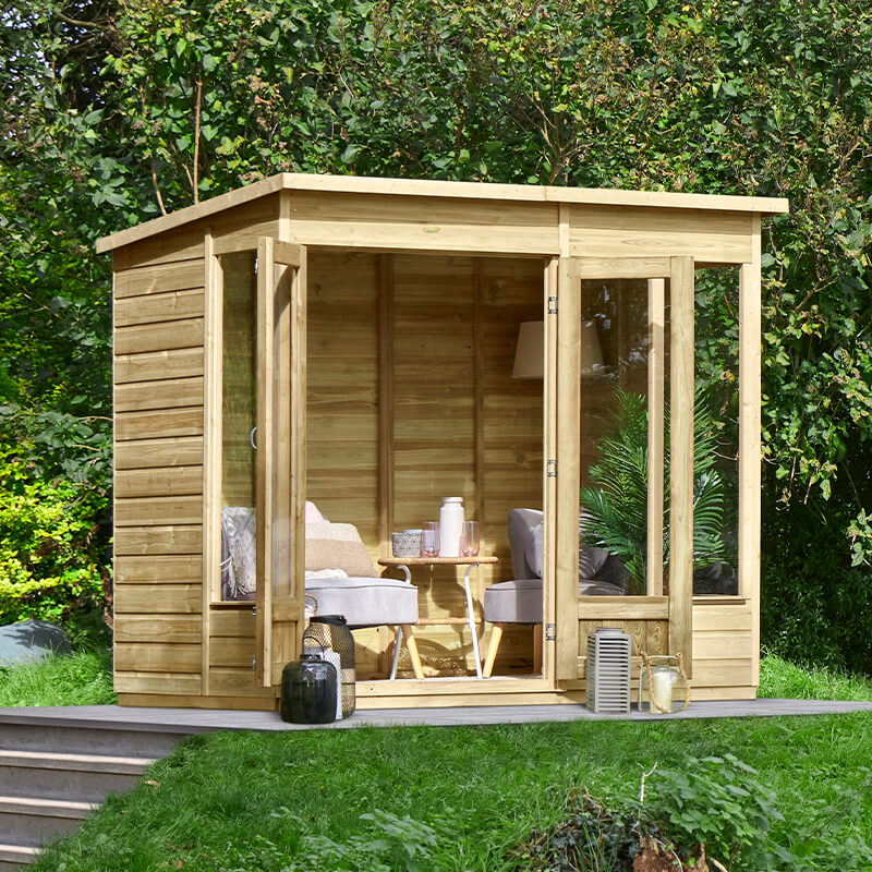 Wooden summer house with a pent roof, featuring double doors, large windows, and a natural wood finish, set in a green garden with outdoor furniture visible inside.