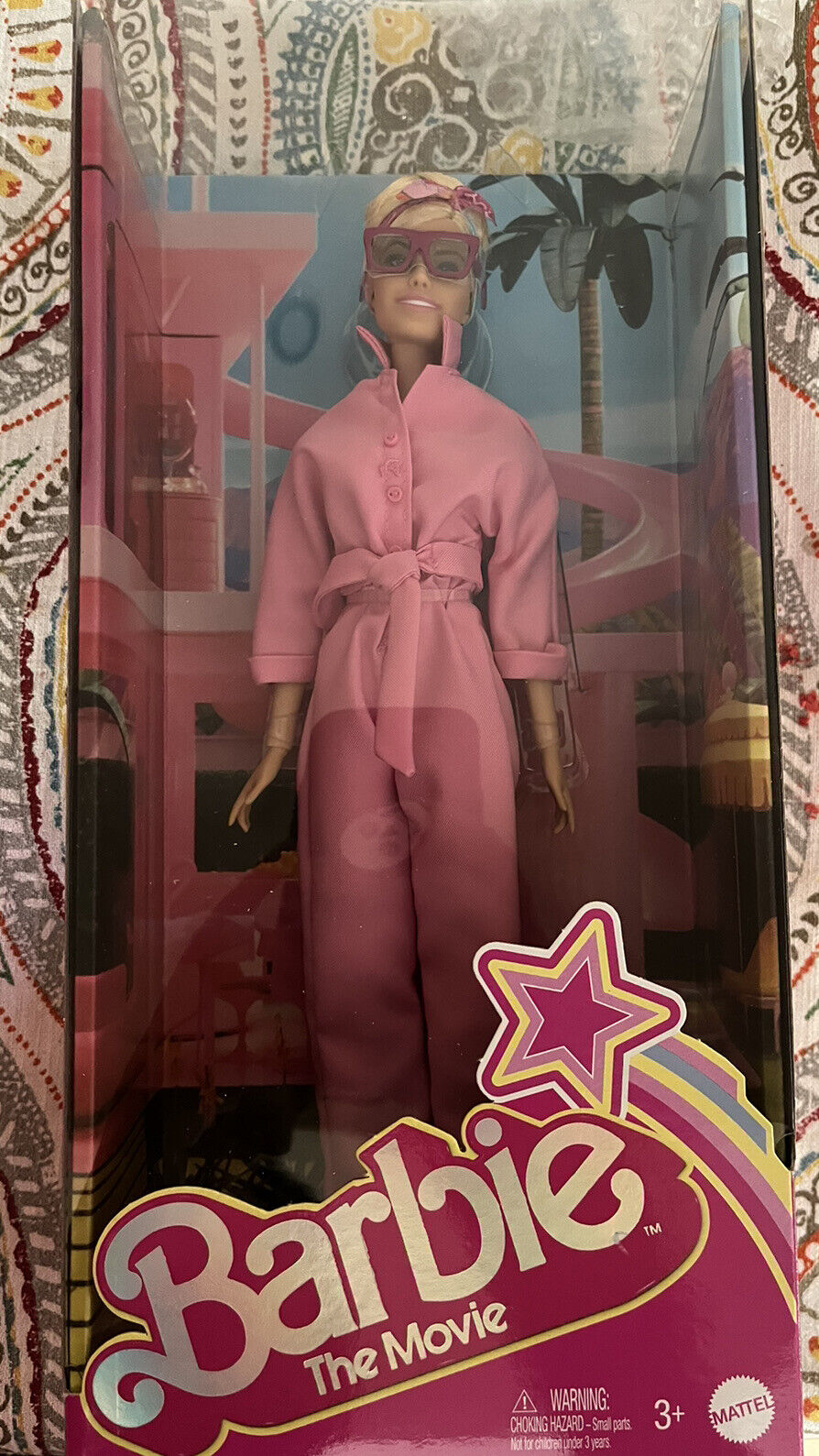 Barbie doll dressed in a pink outfit with a matching robe, wearing stylish glasses, packaged in a colorful box featuring "Barbie The Movie" branding.