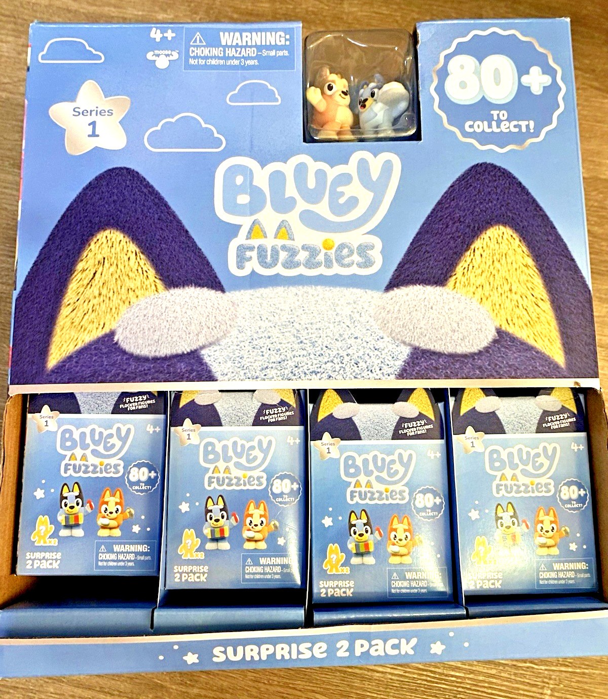 Two-pack mystery figures featuring Bluey characters in a colorful box with a blue background, showcasing various styles and a warning label for ages 4 and up.