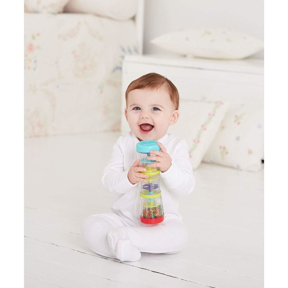 A smiling baby in a white outfit holds a colorful Early Learning Centre Rainmaker Sensory Toy, featuring a transparent body with beads and a blue top, sitting on a light wooden floor.