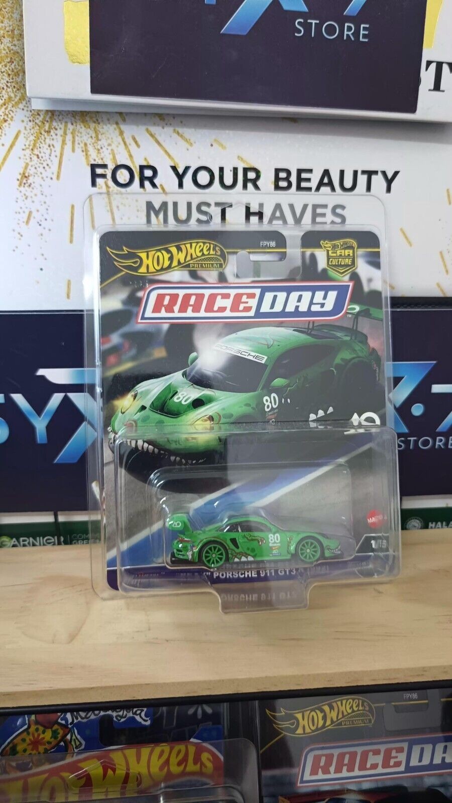 Green Hot Wheels Porsche 911 GT3 R car with dinosaur-themed design, featuring large eyes and teeth, displayed in a clear plastic packaging labeled "Race Day."