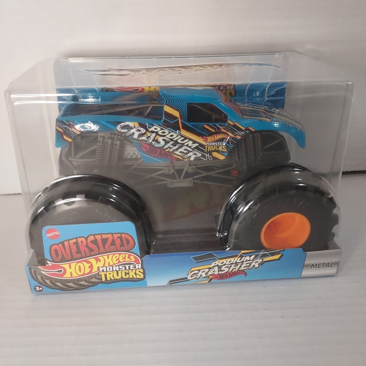 Blue Hot Wheels Monster Trucks Podium Crasher vehicle in a clear plastic box, featuring oversized black wheels with orange rims and colorful graphics on the body.