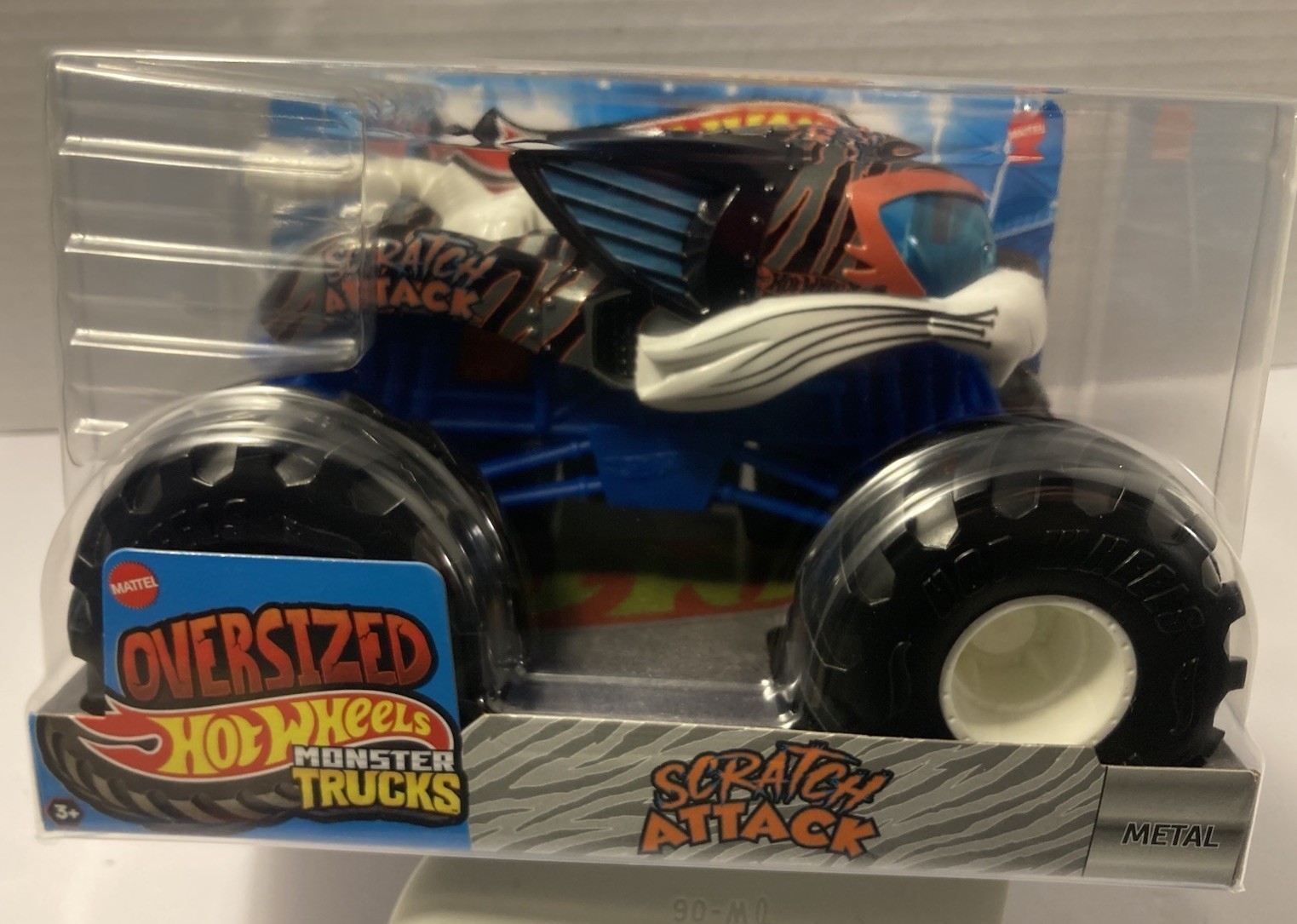Hot Wheels Monster Trucks Scratch Attack Vehicle features a vibrant orange and blue design, oversized black tires, and a stylized cat face on the hood, packaged in a clear plastic box.