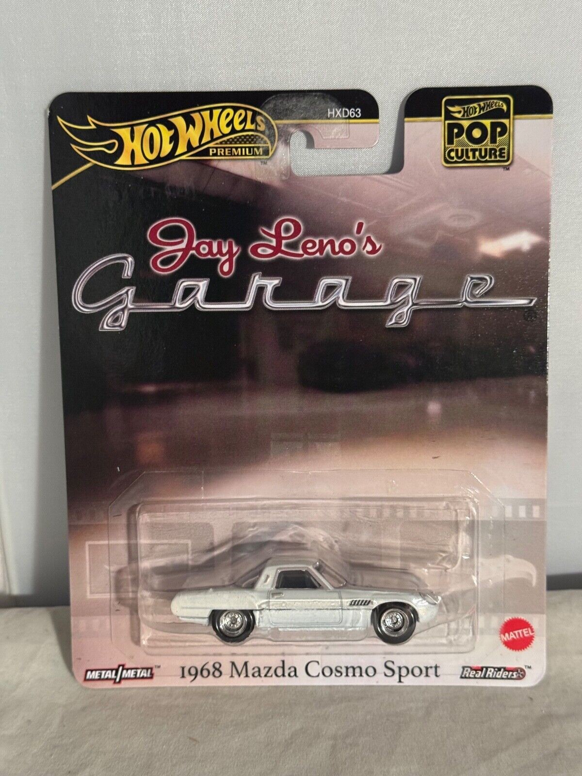 White 1968 Mazda Cosmo Sport Hot Wheels Premium toy car in a clear plastic case, featuring a metallic finish and detailed design, packaged with a Jay Leno's Garage theme.