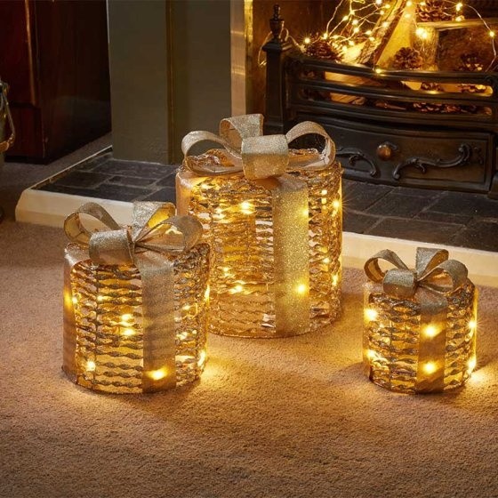 Three glittering gold gift boxes with textured surfaces and large bows, illuminated with warm lights, arranged near a fireplace.