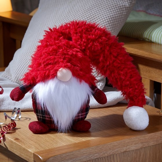 A plush winter gnome with a long, fluffy red hat, white beard, and plaid body, sitting on a wooden surface surrounded by festive decorations.