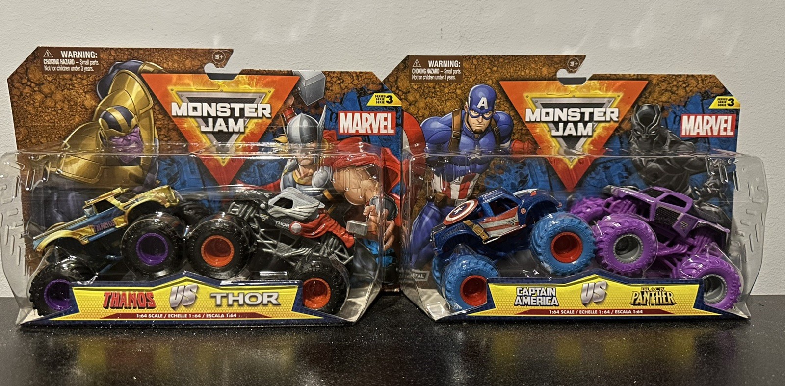 Two die-cast monster trucks in packaging: Thanos in black and gold, Thor in red and silver, alongside Captain America in blue and red, and Black Panther in purple, all 1:64 scale.