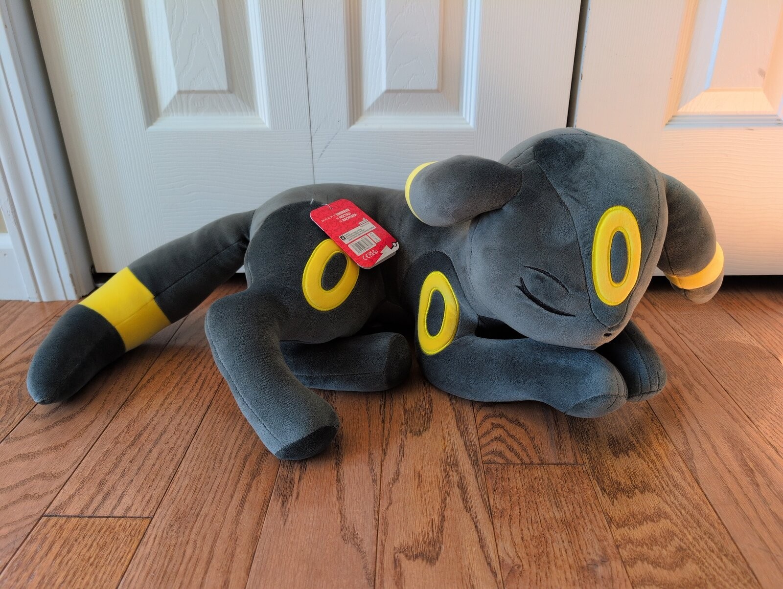Soft plush toy of a sleeping Umbreon, featuring a dark gray body, yellow markings, and large ears, resting on a wooden floor with a red tag attached.