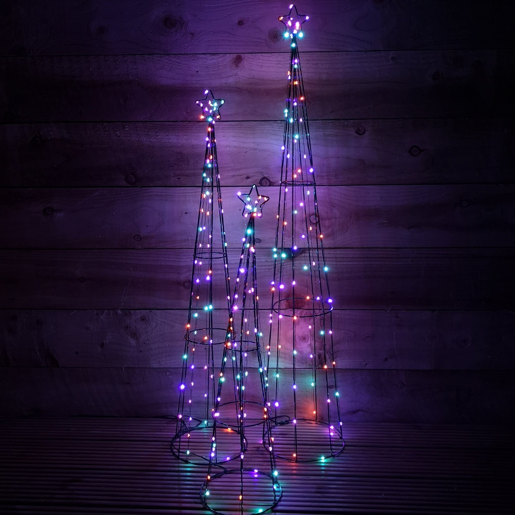 Three wireframe pyramid decorations in varying heights, adorned with colorful LED lights in rainbow hues, featuring star-shaped tops, set against a dark wooden background.