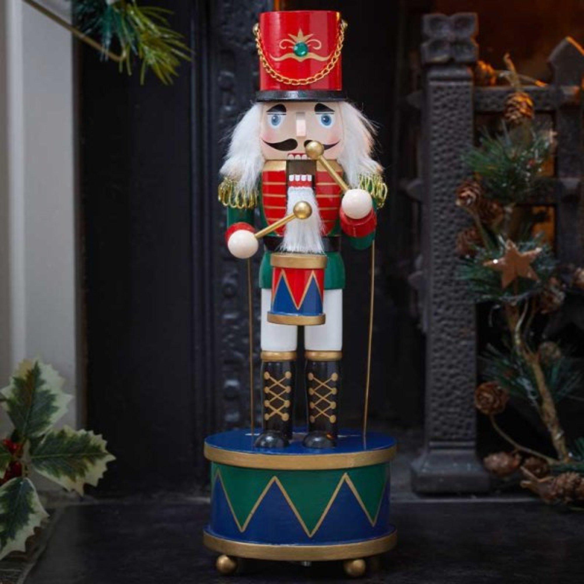 Wooden nutcracker decoration with a green base, featuring a drummer in a red hat, white beard, and colorful uniform, playing a drum with a musical element.