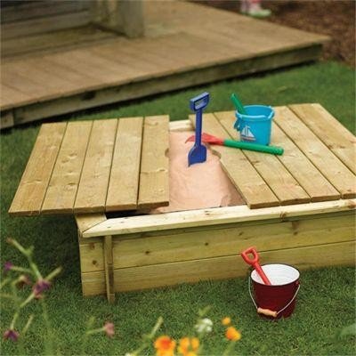 Wooden sandpit made from natural timber, featuring a hinged lid, with colorful plastic shovels and buckets resting on top, set on green grass.