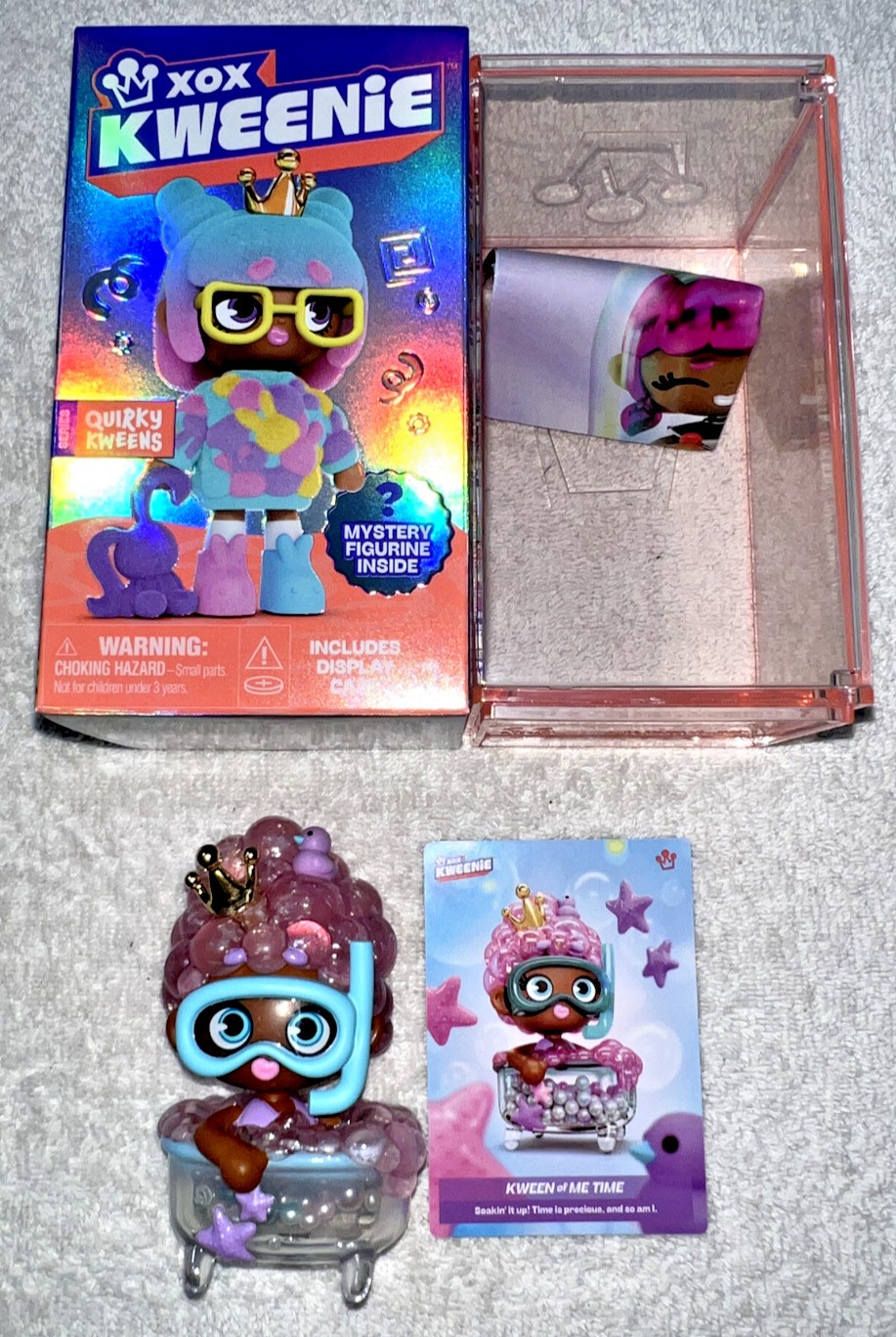 A colorful box labeled "Xox Kweenie" contains a quirky figurine with pink hair, glasses, and a crown, alongside a character card and a clear display case.