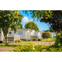 White holiday homes with porches in a green park setting, surrounded by trees and shrubs under a blue sky.