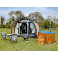 Curved wooden glamping pod with glass doors, surrounded by chairs, a fire pit, and a wooden hot tub on grassy field.