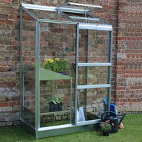 Small silver aluminum-framed greenhouse with clear panels, sloped roof, and sliding door against a brick wall.