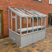 Green-painted wooden lean-to greenhouse with clear panels, sloped roof, and door, set against a brick wall.