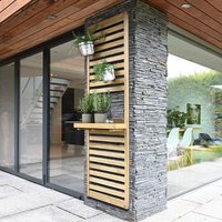 Tall wooden slatted wall planter with one shelf, mounted on stone pillar, holding potted plants.
