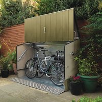 Green metal bike shed with ramp, open doors revealing two bicycles inside, surrounded by plants and wooden fence.