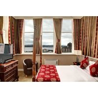 Hotel room with large windows, red and gold curtains, patterned bedspread, armchair, TV, and view of water.