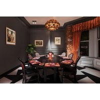 Elegant dining room with dark walls, ornate chandelier, black table set for six, and large windows with orange drapes.