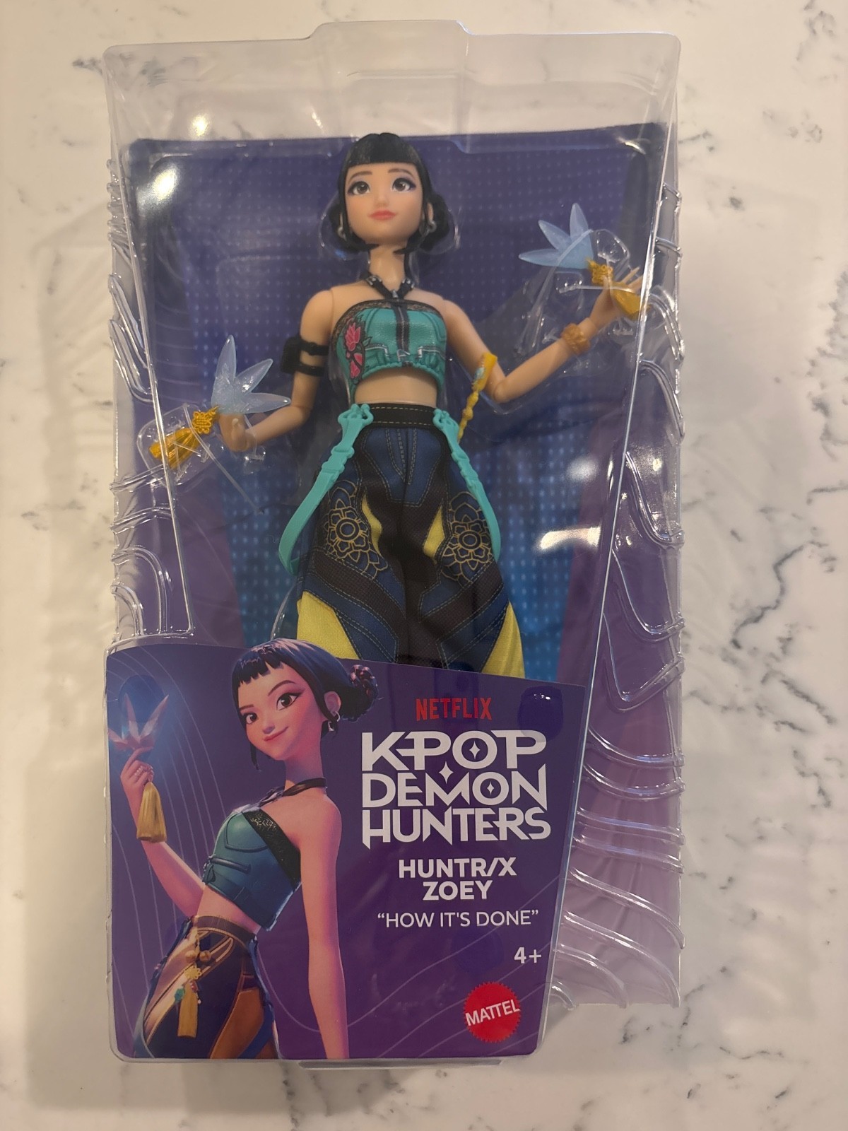 Kpop Demon Hunters Huntr/x Zoey doll features a stylish outfit with blue and black accents, holding translucent blue accessories, packaged in a vibrant box with Netflix branding.