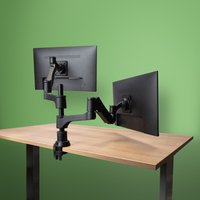 Black dual monitor arm with adjustable joints, mounted on a wooden desk, holding two flat-screen monitors.