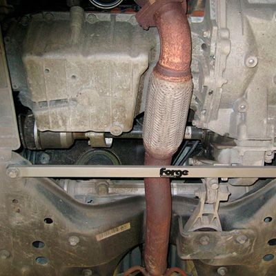 Lower subframe strut brace in stainless steel by Forge, featuring a sleek silver finish, positioned beneath a rusted exhaust pipe and engine components.