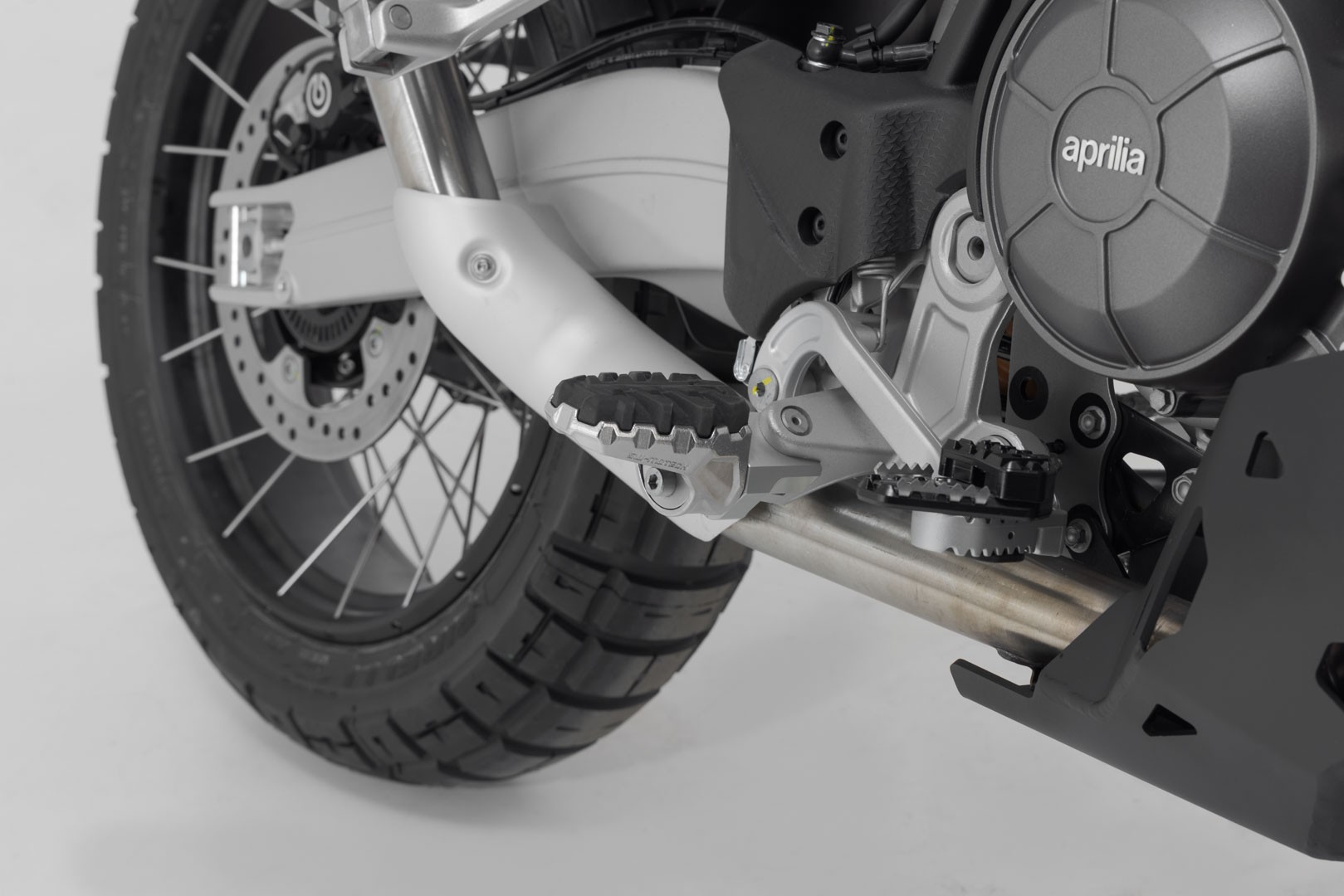 Adjustable SW-Motech Evo footpeg kit in silver and black, featuring a sturdy metal construction with textured rubber grip, mounted on a motorcycle near the rear wheel.