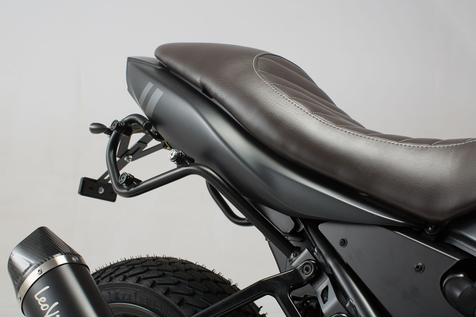 Black metal side carrier for Suzuki SV650, featuring a sleek design, mounted beside a brown leather seat, with a visible exhaust pipe and tire in the background.