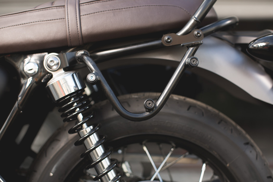 Black metal SLC side carrier for Triumph Bonneville T100/T120, featuring sturdy mounting points and sleek design, positioned near the rear wheel and seat.