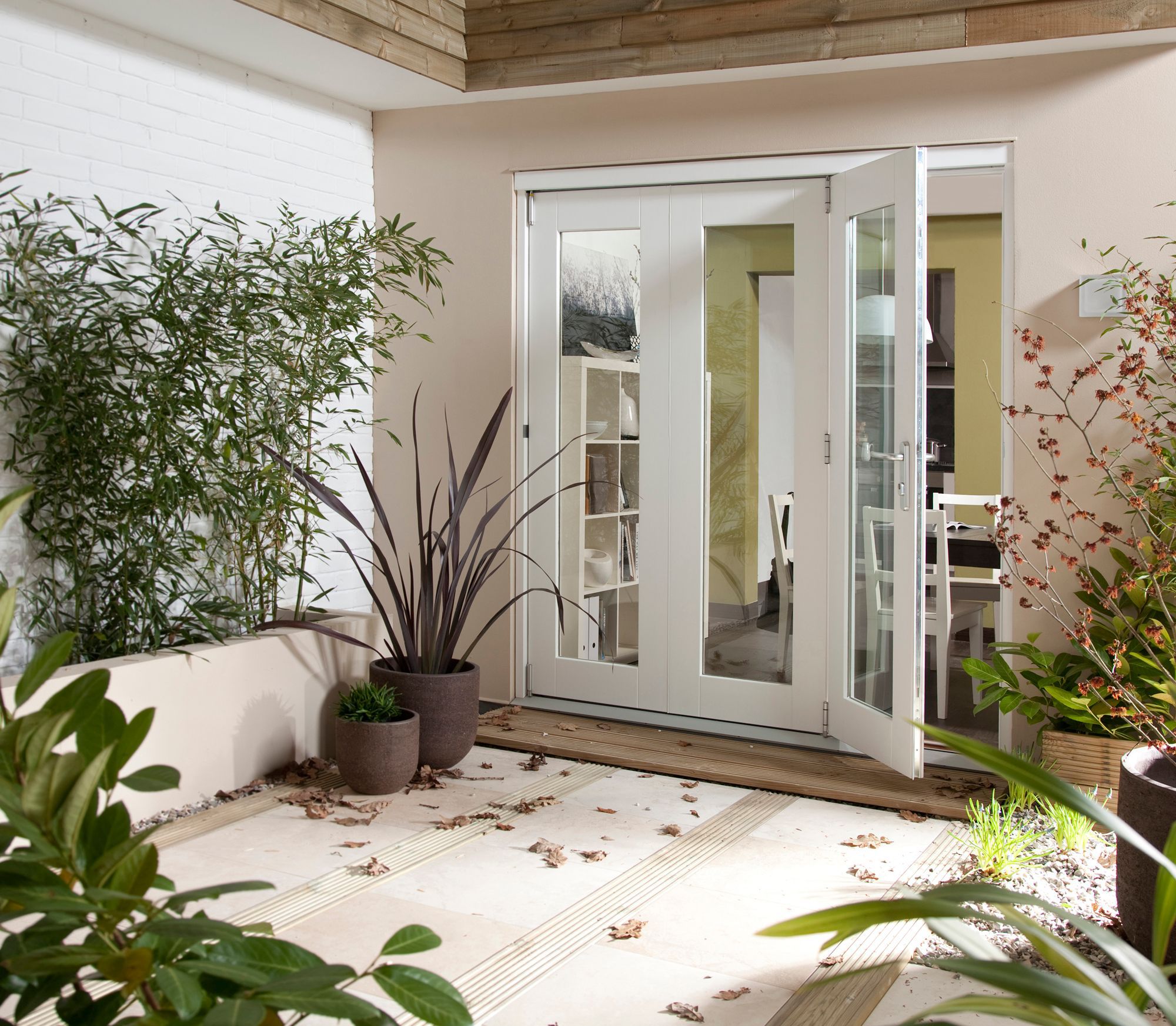 Modern sliding glass doors with a white frame open to a patio area, surrounded by potted plants and bamboo, featuring a light-colored tiled floor and a cozy indoor space visible beyond.