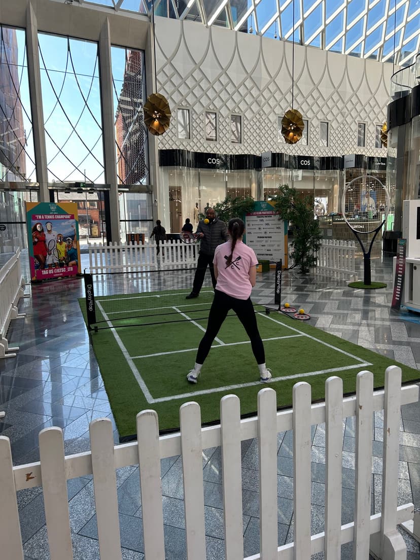A vibrant pop-up tennis court in Victoria Gate, Leeds, with players enjoying the game inside a modern shopping center.