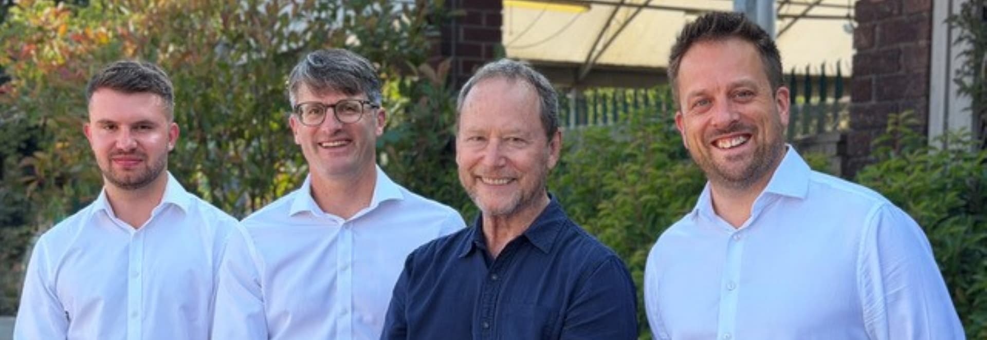 Four smiling men in white shirts standing outdoors with greenery