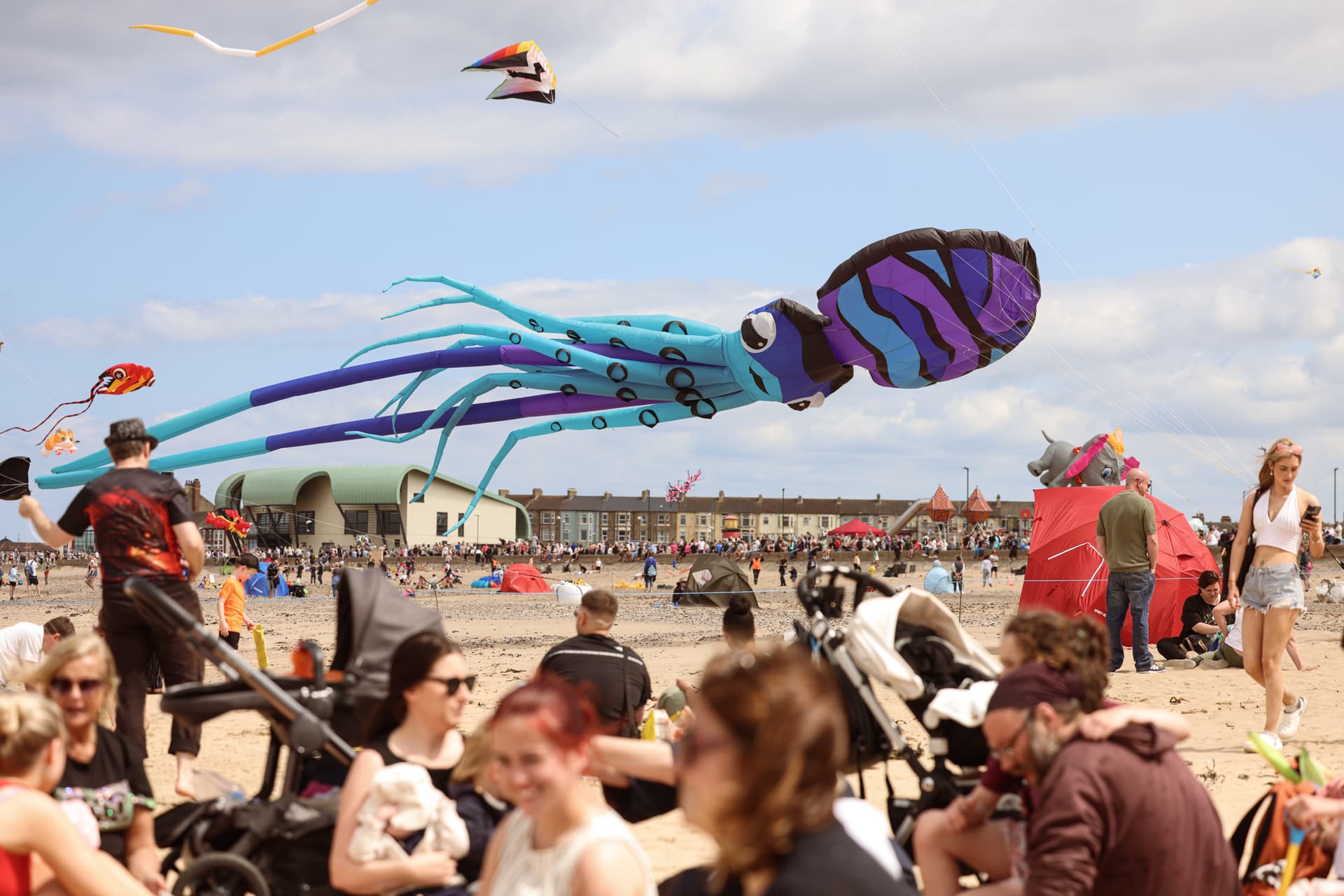 Colorful kites shaped like sea creatures flying over a busy beach