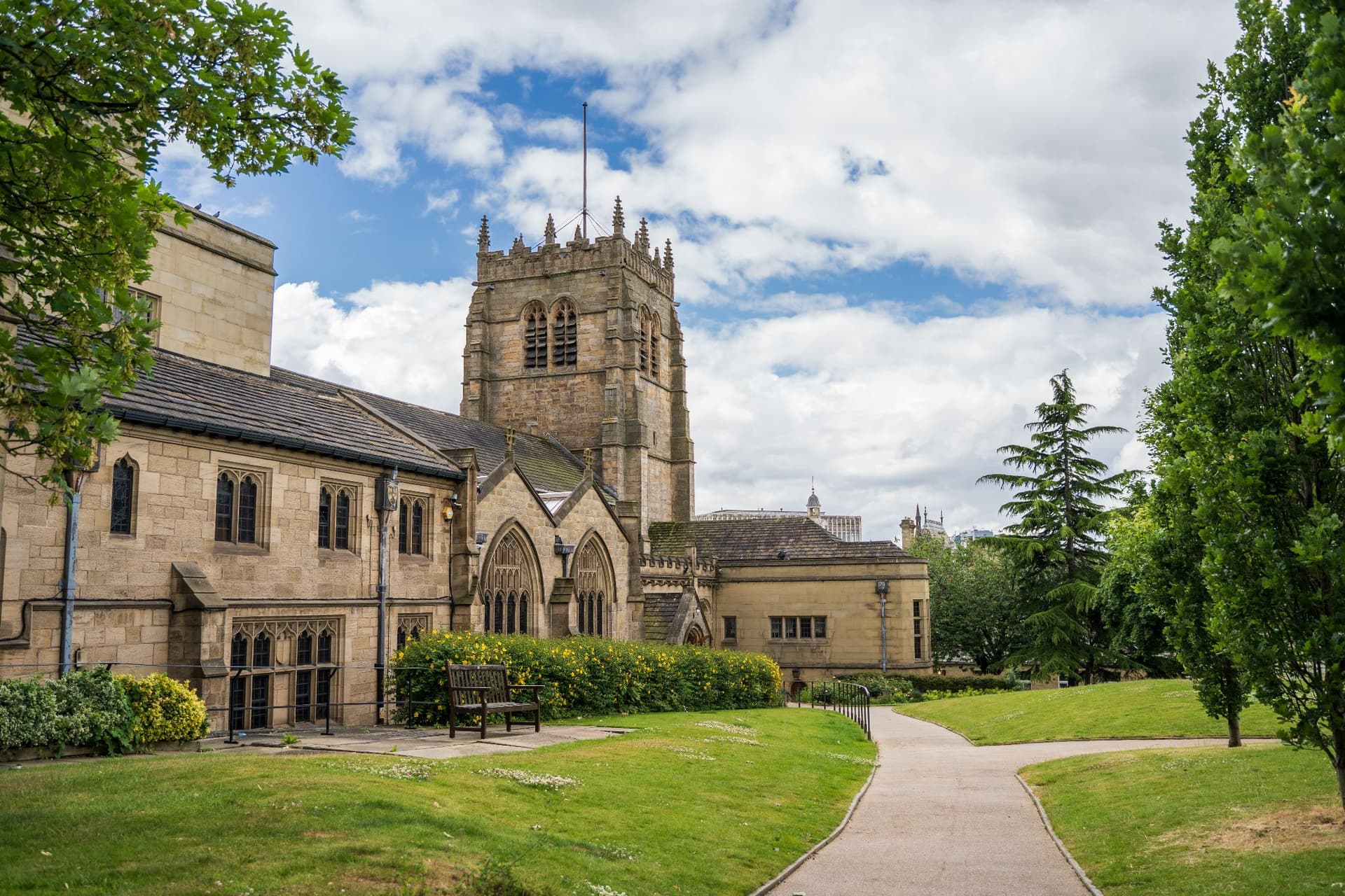 Bradford Cathedral's historic architecture surrounded by lush green gardens and a pathway under a partly cloudy sky.