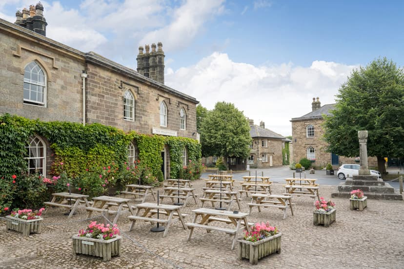 Historic Ripley village square featuring stone buildings, flower-filled planters, and empty picnic tables under a blue sky.