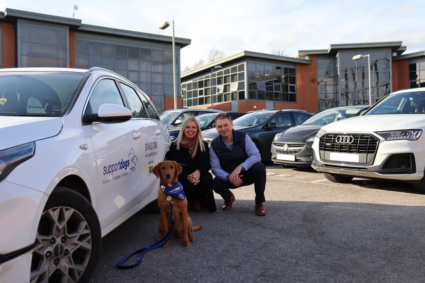 A woman and a man kneel beside a golden retriever puppy in front of a modern building and parked cars in Yorkshire.