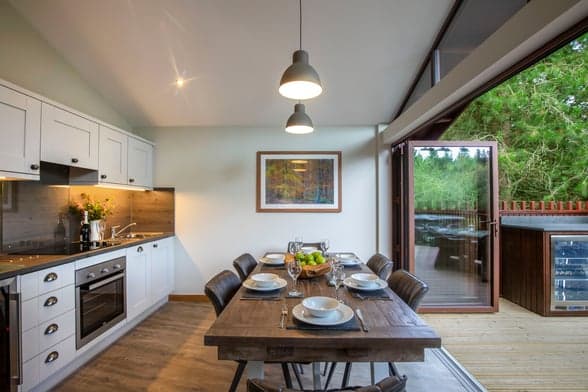 Modern dining area with a wooden table set for six, large windows showcasing greenery, and a stylish kitchen in Yorkshire.