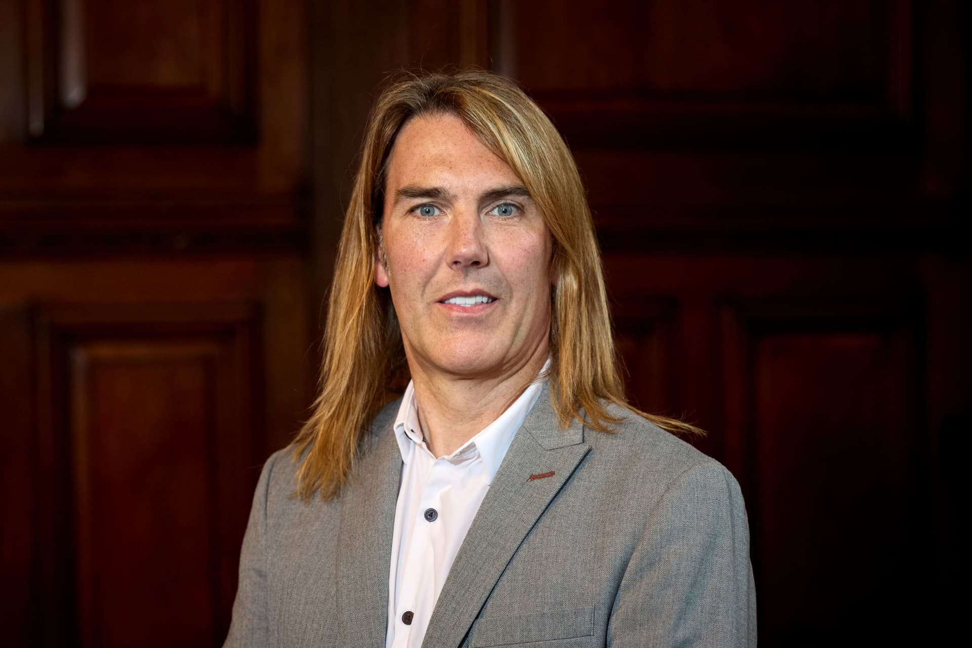 Person in a grey suit with long hair against wooden paneling