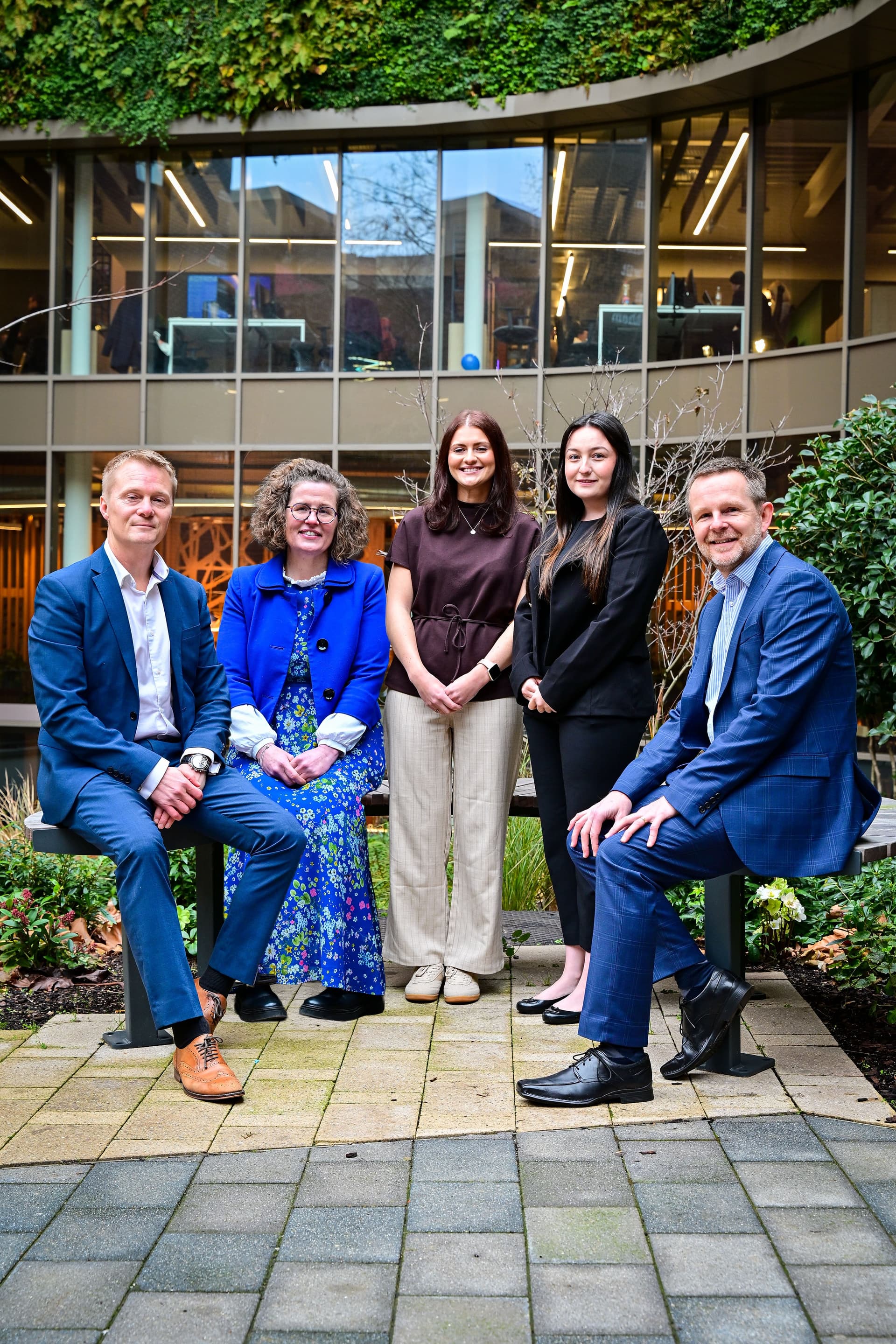Five professionals pose together in a modern courtyard, surrounded by greenery and contemporary architecture, smiling confidently.